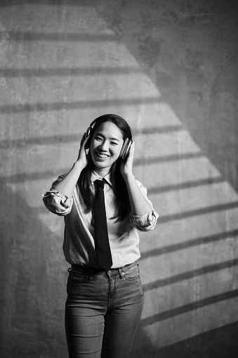 Portrait of a beautiful modern Japanese female listening music on wireless headphones in studio. Black and white.