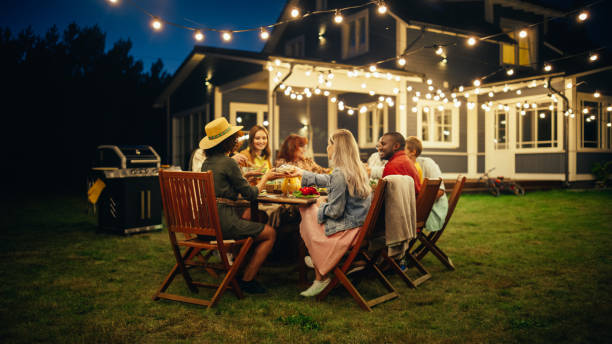 familiares y amigos multiculturales celebrando afuera en casa por la noche. grupo de niños, adultos y ancianos se reunieron en una mesa, teniendo conversaciones divertidas. comer barbacoa y verduras. - front or back yard house family barbecue fotografías e imágenes de stock