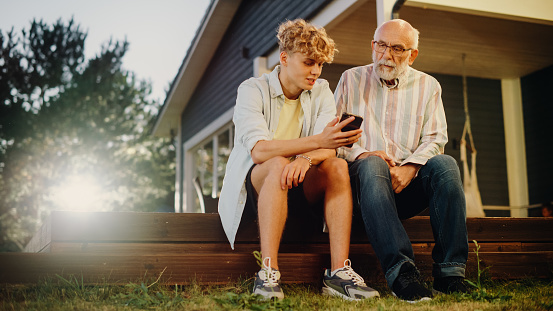 Handsome Teenage Grandson Teaching His Grandfather to Use a Smartphone. Young Man Showing Family Photos and Videos to His Grandparent. Relatives Sitting Outside on a Porch on a Nice Summer Day.