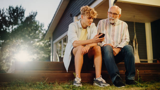 handsome teenage grandson teaching his grandfather to use a smartphone. young man showing family photos and videos to his grandparent. relatives sitting outside on a porch on a nice summer day. - senior adult technology child internet imagens e fotografias de stock