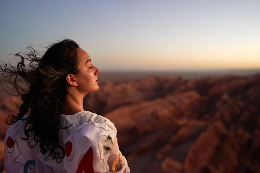 rear view young latin woman standing at the viewpoint over the Valle de la Luna or valley of the moon at sunset with eyes closed enjoying the wind