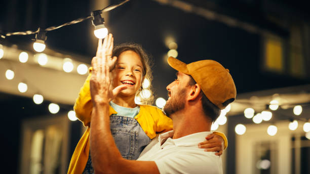 orgulloso y guapo padre ayudando a su pequeña y hermosa hija a cambiar una bombilla en la instalación del patio trasero de fairy lights en casa. padre e hija chocan los cinco y celebran la solución exitosa. - fiesta en el jardín fotografías e imágenes de stock