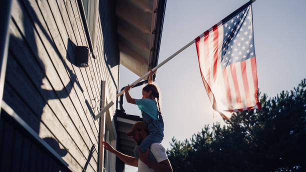 Proud Patriotic Dad Holding His Young Daughter of His Shoulders, Helping Her to Put the United States of America Flag on the Wall of Their House to Celebrate a National Holiday. Sunny Day in USA. stock photo