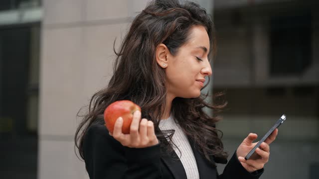 Businesswoman biting an apple while holding smartphone and looking at the screen. Young lady standing outdoor and using mobile phone. Lifestyle, food concept. Real time