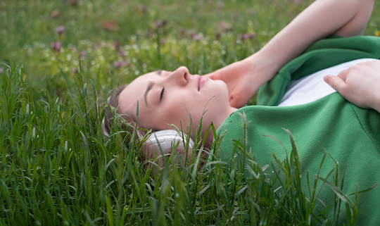 A teenage girl listens to music on headphones, lies on the green grass in the park, smiles and shows her tongue in a white t-shirt and a green sweater thrown over, summer mood as a lifestyle