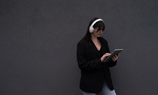A beautiful girl in white headphones and a black jacket holds a tablet outside, work outside, listen to music and sing along, office worker
