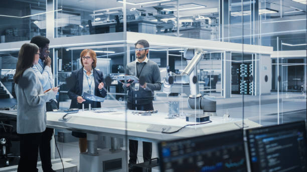 istock Diverse Team of Industrial Robotics Engineers Gathered Around a Table With Robotic Arm. Scientists Use Tablet Computer to Manipulate and Program the Robot to Pick Up and Move a Metal Component. 1481683923