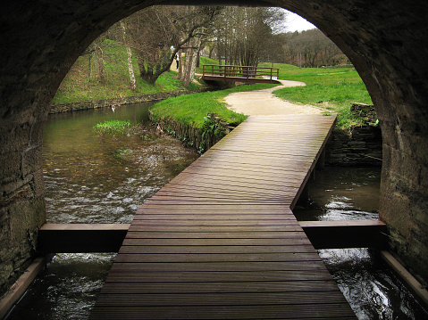 Wooden footbridge and boardwalk under a tunnel. Hiking trail, river and landscape in the background. Rato river, Lugo, Galicia, Spain.
