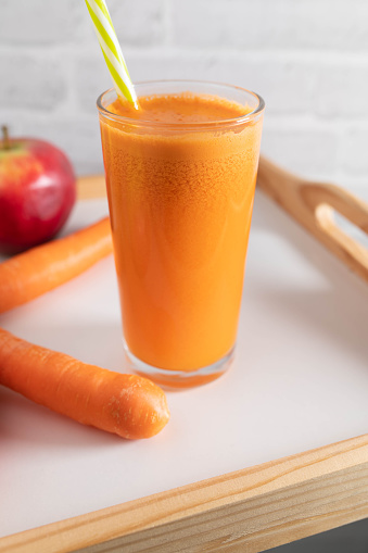 Homemade freshly squeezed carrot, apples juice or smoothie. Served in a drinking glass with straw on a white wooden tray. Closeup, front view with copy space