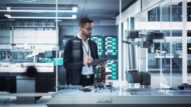 istock Industrial Robotics Professional Interacting with Robotic Arm During a Research Phase in a High Tech Startup. Scientist Uses Tablet Computer to Manipulate and Program the Robot to Move a Microchip. 1481702016