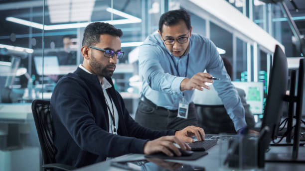 istock Male Project Supervisor Talks to an Industry 4 Engineer Who Works on Desktop Computer in Robotics Startup Industrial Office. Software Developer Discussing Work Related Tasks During a Meeting. 1481702073