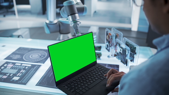 Over the Shoulder Shot of a Robotics Engineer Using a Laptop Computer with Green Screen Mock Up Display Screen. Robotic Arm Holding a Microchip. Automation Startup Research and Development Office.