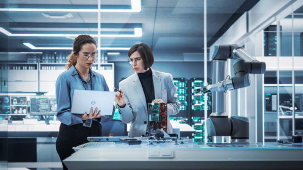 istock Two Female Working in High Tech Facility. Robotics Engineer and Machinery Operator Collaborate on a Robot Arm Project, Using a Laptop Computer to Operate the Robot Arm in Technology Startup Lab. 1481703572