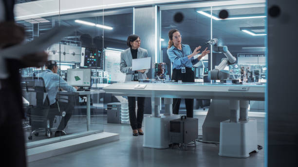 istock Two Female Working in High Tech Facility. Robotics Engineer and Machinery Operator Collaborate on a Robot Arm Project, Using a Laptop Computer to Operate the Robot Arm to Move Around a Microchip. 1481703615