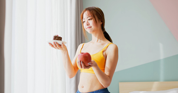 asian diet woman standing in bedroom is making choices about food - she holding apple and cake