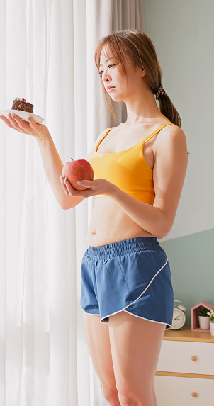 asian diet woman standing in bedroom is making choices about food - she holding apple and cake