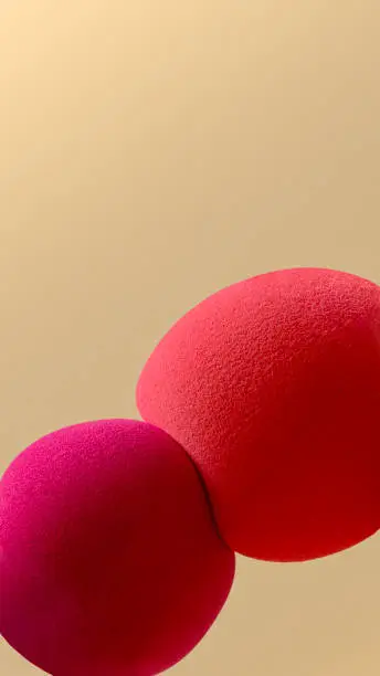 Macro shot of two spherical makeup sponges - red and orange, squeezing against beige background.