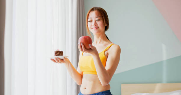 asian diet woman standing in bedroom is making choices about food - she holding apple and cake
