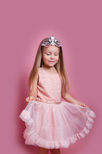 Cute child girl in elegant pink dress and tiara celebrates birthday party on pink studio background.