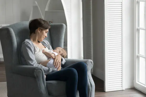 Photo of Happy pretty new mom breastfeeding little baby, resting in armchair