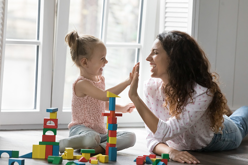 Cheerful mom and kid girl clapping hands, giving high five over colorful toy towers stacked from wooden constructing blocks, playing on heating floor, celebrating successful building