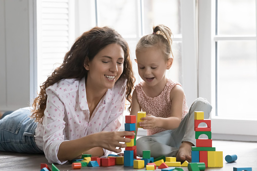 Happy mom and cheerful cute preschool kid girl playing on heating floor at home, stacking colorful cubes, constructing toy towers from building blocks, talking, laughing, smiling, enjoying leisure