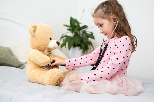Cute little Caucasian girl with a stethoscope and a toy bear is playing doctor at home sitting on the bed. Child treats a toy bear