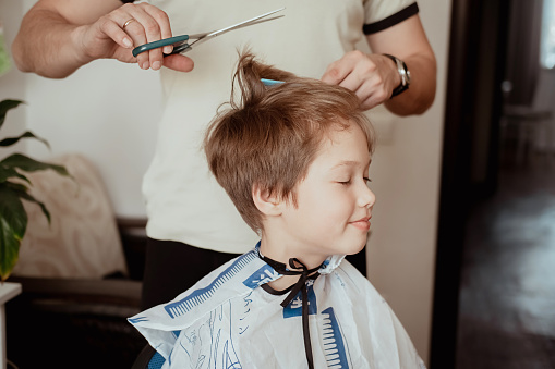 Hairdressing salon at home in quarantine conditions. Father cuts son's hair with scissors. Man cuts boy's hair at home. Concept of economy in an economic crisis