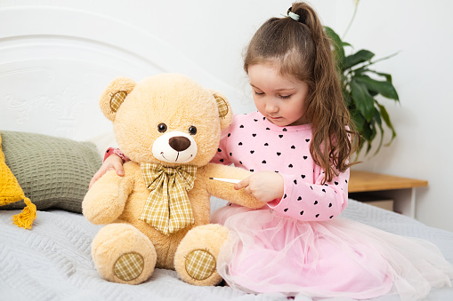 Cute little Caucasian girl is sitting on bed and measuring temperature of toy bear with an electronic thermometer. Child plays doctor at home
