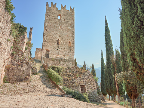 Low-angle view of the Torre Grande, the main tower of the Castle of Arco di Trento (Castello di Arco), a medieval fortress located on a prominent spur high above the town of Arco, a few steps from Lake Garda. The warm, bright light of a spring afternoon, a perfectly clear sky, a paved walkway crossing the ruins of the castle, lush vegetation and cypress trees. High level of detail, natural rendition, realistic feel. Developed from RAW.