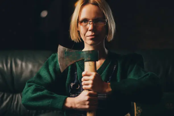 Photo of A woman with a serious expression holds an ax in her hands while sitting on a sofa. The woman is on the warpath. A strong, strong-willed, determined woman of middle age.
