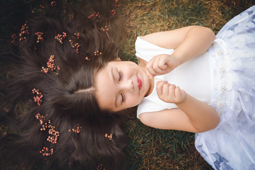 Little girl in white dress lying on the grass, she is smiling, her hair is very long and princess-like, she plays with her hands on the theme of children's day.