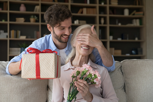 Cheerful adult son guy giving surprise gift to old senior mom, covering woman eyes with hand, holding present box, flowers bouquet, celebrating international women day, birthday, mothers day