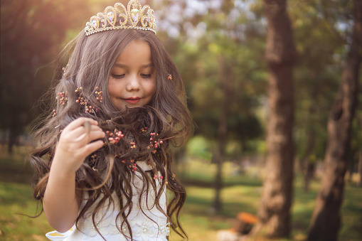 Little girl in white dress and princess crown, she plays with her long blonde hair, copy space, children's day theme.