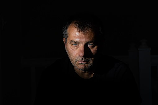 A striking portrait captured on a black backdrop with distinct shadows, featuring a male model in his fifties from the Balkan region who is staring directly at the camera with an unshaven face.