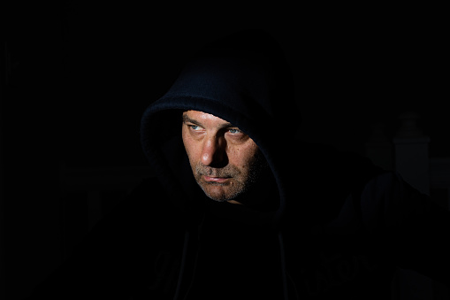 In a dimly lit room, a man in his fifties with stubbly, unshaven facial hair is wearing a hood that is illuminated by a harsh light.