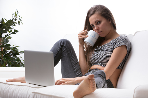 A young woman is sitting on the couch drinking something warm from her mug in front of her laptop. She is barefoot and relaxed. Her gaze is challenging and serious, yet attractive and sexy. She is sel