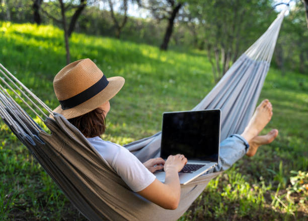 Young woman in a hat using a laptop while lying in a hammock in a summer garden. Summer lifestyle stock photo