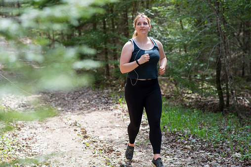Young woman wearing all black fitness clothing and earphones for listening to music is jogging through the tranquil nature trail in a forest to de-stress, exercise, and appreciate nature.