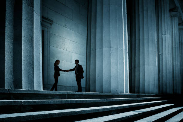 Man And Woman Shake Hands In The Shadows A man and woman shake hands in the shadow of a portico behind a row of tall columns. corruption stock pictures, royalty-free photos & images