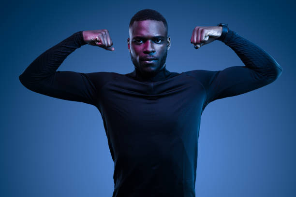 Self assured young bearded African American male athlete in stylish black activewear showing biceps and looking at camera against blue background