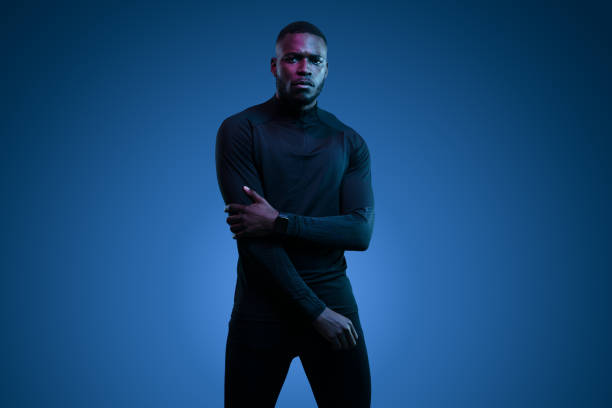 Thoughtful young brutal African American male model with dark hair and beard in total black activewear with partial crossed arms looking at camera against blue background