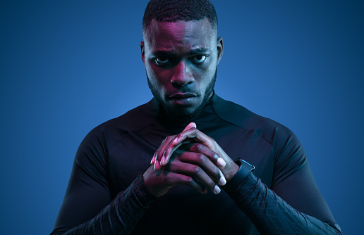 Determined adult bearded African American Athlete in black sportive turtleneck standing with folded arms in dark room with neon light and looking at camera