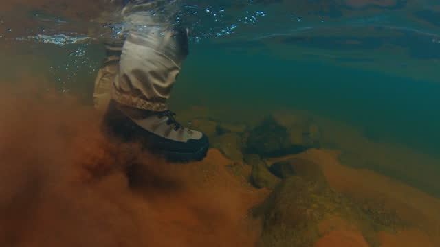 Angler walks in the river underwater view. Fisherman wearing fishing boots and waders walks on the sandy bottom of the river