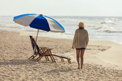 A girl walks along the waters edge of Perdido Key Beach on a windy day. With  lounge chairs and umbrella in the background.