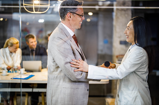 Happy businessman shaking hands with Asian female doctor in the office. The view is through glass. There are people in the background.