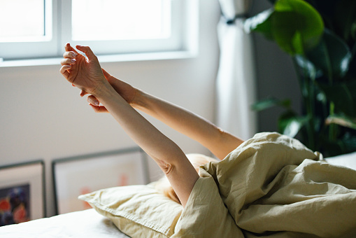 Unrecognizable woman laying in bed in the morning. She is stretching her arms above her.