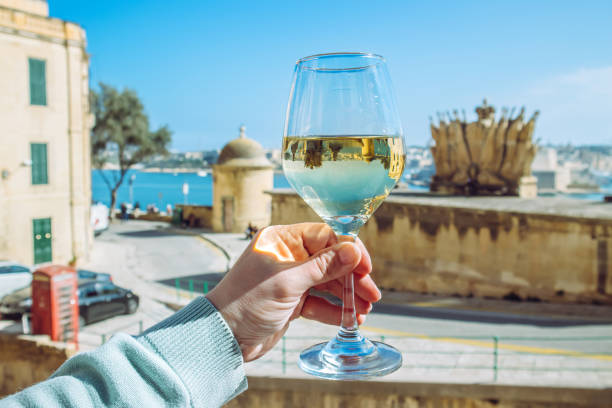 Selective focus on woman hand holding glass of white wine in beautiful city of Valletta in Malta. Background with blue sky and sea on sunny day. Wine tour concept. stock photo