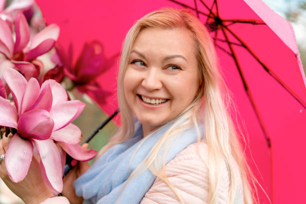 blonde frau im blauen mantel, die am frühlingstag in einem park zwischen blumen spazieren geht - magnolia blossom flower head single flower stock-fotos und bilder