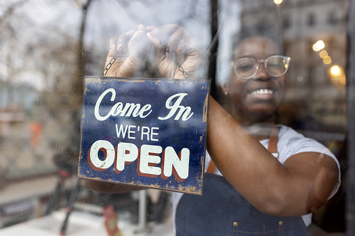 African woman hanging an open sign on the glass door of zero waste grocery store. Female store assistant put on open sign board on front door.